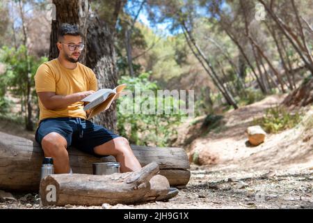 Boy sitting on a log in a yellow T-shirt and glasses reading a book in the middle of nature surrounded by trees. Stock Photo