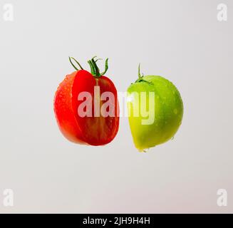 Red, ripe, fresh and tasty tomato with drops and green, unripe tomato cut in half. Minimal food levitation concept. Abstract composition. Stock Photo