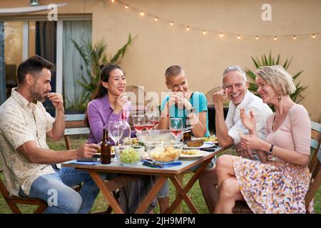 Group of happy friends having fun drinking red wine at barbecue picnic garden party. Middle aged people eating a tasty meal in the backyard. Stock Photo