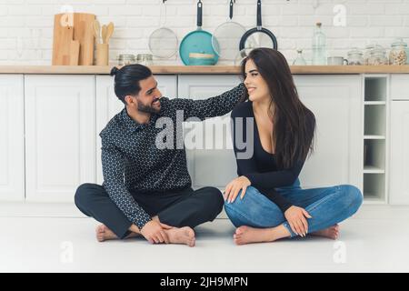 Closeness and tenderness. Barefoot heterosexual couple - bearded formally dressed handsome Cuban man and long-haired caucasian casually dressed woman - sitting on the white kitchen floor. Man touching his young wife's hair. Modern kitchen interior. High quality photo Stock Photo