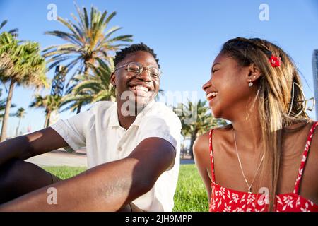 Cheerful young African couple laughing and having fun on their first date in the park in a romantic outdoor lovers scene, sitting on the grass talking Stock Photo