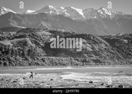 Kaikoura New Zealand - May 11 2022; Stony Mangamaunu, beach near Kaikoura with surf rolling in and surfers under backdrop of coastal range with snow c Stock Photo