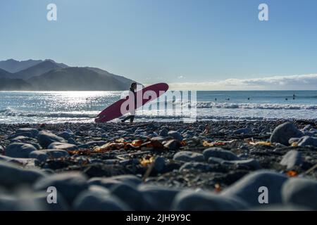 Kaikoura New Zealand - May 11 2022; Stony Mangamaunu, beach near Kaikoura with surf rolling in silhouette and young woman surfer carrying board across Stock Photo