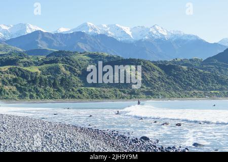 Kaikoura New Zealand - May 11 2022; Stony Mangamaunu, beach near Kaikoura with surf rolling in and surfers under backdrop of coastal range with snow c Stock Photo