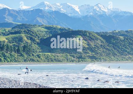 Kaikoura New Zealand - May 11 2022; Stony Mangamaunu, beach near Kaikoura with surf rolling in and surfers under backdrop of coastal range with snow c Stock Photo