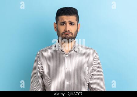 Bearded upset businessman standing and looking at camera with dissatisfied sadness face, expressing sorrow, having bad mood, wearing striped shirt. Indoor studio shot isolated on blue background. Stock Photo