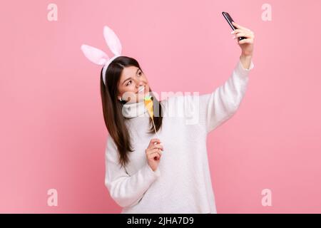 Side view portrait of funny girl with bunny ears streaming or has video call with carrot in hands, wearing white casual style sweater. Indoor studio shot isolated on pink background. Stock Photo