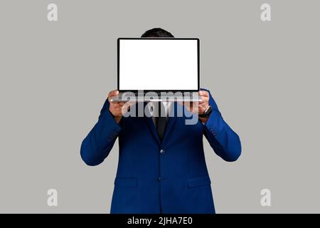 Anonymous unknown man hiding his face behind laptop with white blank display with space for advertisement, wearing official style suit. Indoor studio shot isolated on gray background. Stock Photo