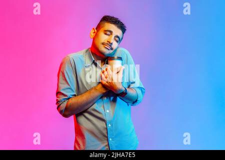 Portrait of tired man in shirt holding coffee cup and sleeping, feeling exhausted bored after stressful working day. Indoor studio shot isolated on colorful neon light background. Stock Photo
