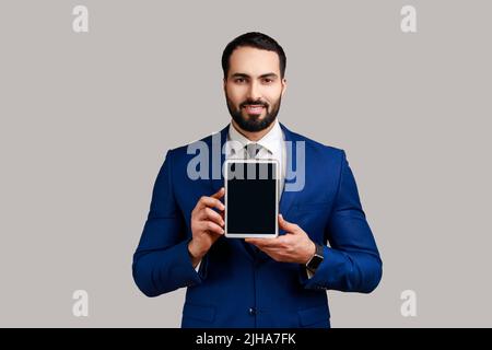Man holding and showing tablet with blank screen with toothy smiling and satisfied face, copy space for advertisement, wearing official style suit. Indoor studio shot isolated on gray background. Stock Photo