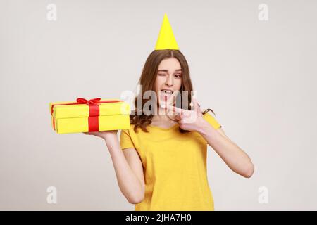 Cute amazed teenage girl with party cone in yellow T-shirt pointing at gift box and winking to camera, showing awesome birthday present. Indoor studio shot isolated on gray background. Stock Photo