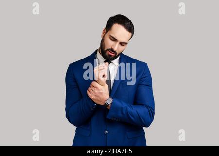 Portrait of businessman standing with grimace of pain, massaging sore wrist, suffering hand injury or sprain, wearing official style suit. Indoor studio shot isolated on gray background. Stock Photo