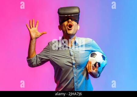 Portrait of excited man in shirt wearing virtual reality glasses and holding soccer ball, raised arm, supporting favorite football tram. Indoor studio shot isolated on colorful neon light background. Stock Photo