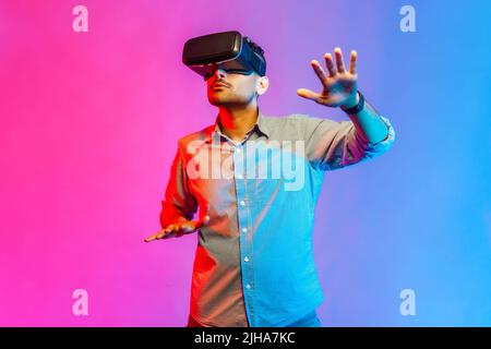 Portrait of concentrated man in shirt discovering new technologies wearing virtual reality headset, futuristic 3d vision. Indoor studio shot isolated on colorful neon light background. Stock Photo