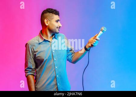 Side view of attractive man in shirt posing with microphone in hands, offers mic, journalist asking questions, looking away with smile. Indoor studio shot isolated on colorful neon light background. Stock Photo