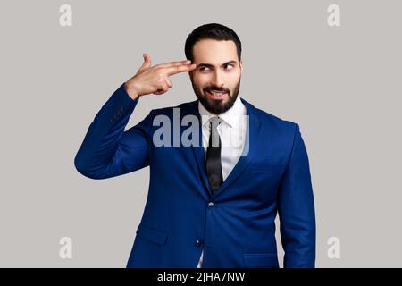 Portrait of bearded man holding fingers near head imitating gun, looking at camera with panic stressed expression, wearing official style suit. Indoor studio shot isolated on gray background. Stock Photo