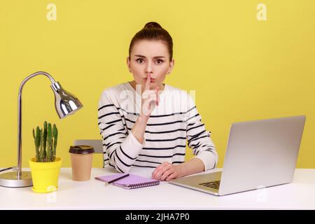 Be quiet. Serious woman in striped shirt sitting at workplace showing silence sign, looking at camera with finger on lips, asking to be silence. Indoor studio studio shot isolated on yellow background Stock Photo