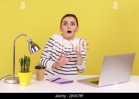 Frightened woman with shocked expression raising hands in stop gesture, defending herself, freaked out of troubles working on laptop. Indoor studio studio shot isolated on yellow background. Stock Photo