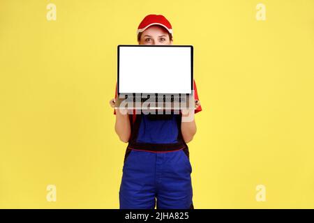 Full length portrait of unknown worker woman standing covering half of face with laptop computer with blank screen for promotional text. Indoor studio shot isolated on yellow background. Stock Photo