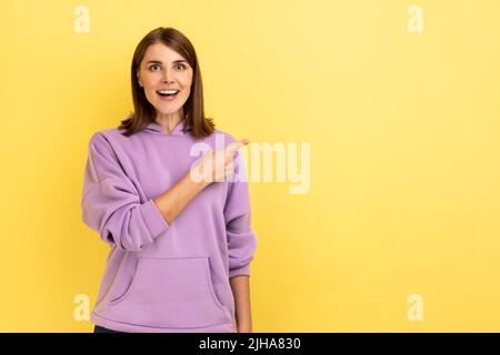 Portrait of optimistic beautiful young woman standing and pointing at copyspace with happy positive face, wearing purple hoodie. Indoor studio shot isolated on yellow background. Stock Photo