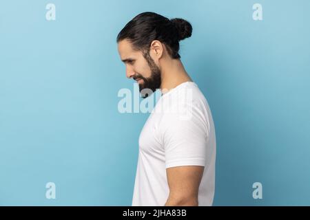 Portrait of sad upset man bodybuilder wearing white T-shirt standing looking down, being in bad mood, suffering depression, expressing sorrow. Indoor studio shot isolated on blue background. Stock Photo