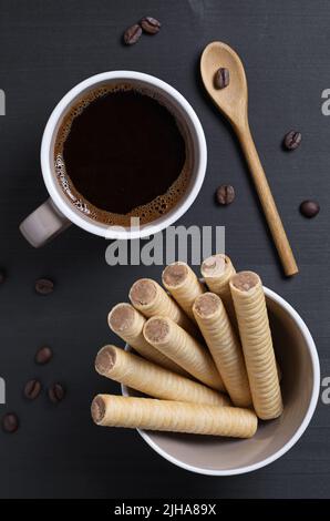Chocolate waffle rolls in bowl and cup of coffee on black wooden table, top view Stock Photo