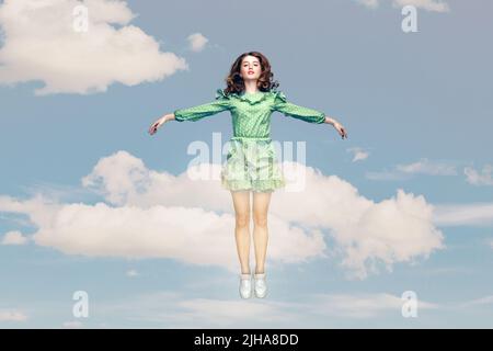 Full length happy calm pretty girl in ruffle dress levitating hovering in mid-air with raised hands as wings, jumping trampoline or flying up in the sky. collage composition on day cloudy blue sky Stock Photo