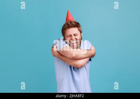 Portrait of extremely happy bearded man wearing party cone hugging himself with excited facial expressing, celebrating his birthday. Indoor studio shot isolated on blue background. Stock Photo
