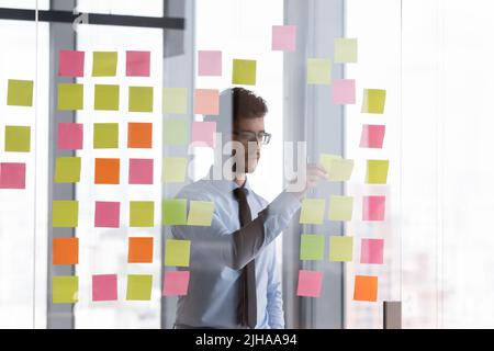 Focused millennial businessman sticking paper notes on glass wall Stock Photo