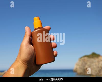 male hand holding up a spray bottle with sunscreen lotion by the sea, important sun protection on vacation Stock Photo