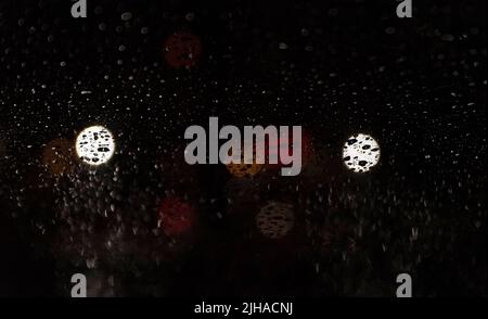 Blurred lights behind car window with water droplets. Raindrops on the windshield of the car at night. Stock Photo