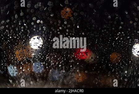 Blurred lights behind car window with water droplets. Raindrops on the windshield of the car at night. Stock Photo