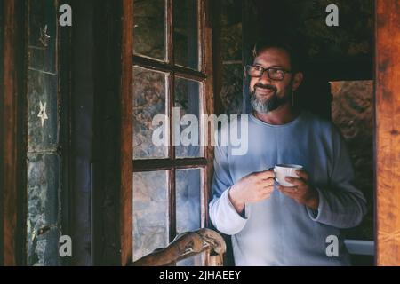 One mature man at home smile and enjoy indoor relax drinking tea or coffee and looking outside the window. People in winter chalet vacation room. Hand Stock Photo