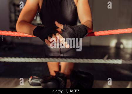 Female boxer leaning on boxing ring ropes with her hands wrapped in black. Unrecognisable young woman training at a boxing gym. Stock Photo