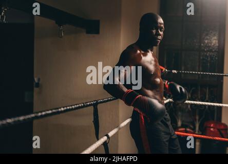 Muscular African boxer looking at the camera while resting in his corner of the boxing ring. Athletic young man having a training session in a boxing Stock Photo