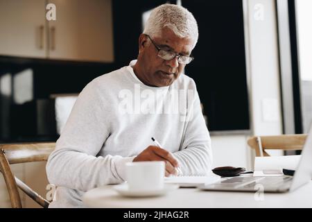 Senior businessman writing in a notebook while sitting at a desk in his home. Mature businessman making plans in his journal while doing remote freela Stock Photo