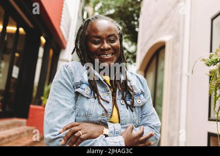 Black woman with dreadlocks smiling happily while standing outdoors. Cheerful senior woman going out in casual clothing. Stock Photo