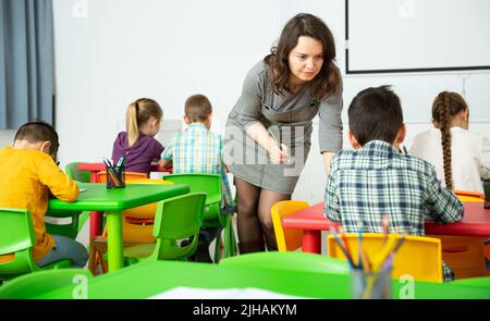 Positive boys and girls studying and female teacher helping in classroom Stock Photo