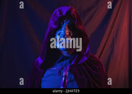 Portrait of a zombie dressed in a shirt and hooded cape facing the camera. The scene is dark, illuminated by blue and orange lights. Stock Photo