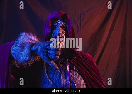 Portrait of a zombie dressed in a shirt and hooded cape making the thumbs-down dislike gesture. The scene is dark, illuminated by blue and orange ligh Stock Photo