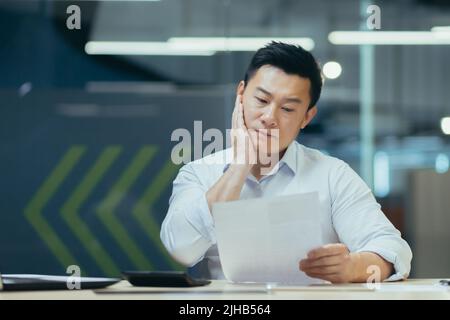 Portrait of a worried young businessman, accountant, financier. He sits at a desk in a modern office over documents, holds his head in his hand, does not know what to do Stock Photo