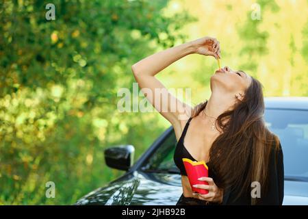 Young woman leaned against the hood of a car on the side of the road and eats french fries Stock Photo