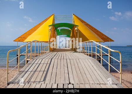 Swimming area or pier on the sea. A large pier with a roof, wooden planks and rusty stairs descending into the water. Bathing and resting place. Stock Photo