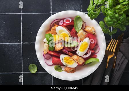 Tuscan Panzanella salad. Fresh tomatoes salad with basil leaves, eggs, olive oil and onion in white bowl on old black background. Vegetarian panzanell Stock Photo