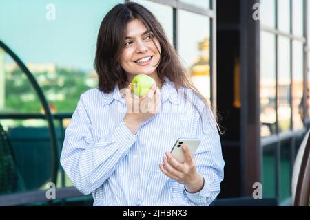 A young woman eats an apple with a smartphone in her hands. Stock Photo