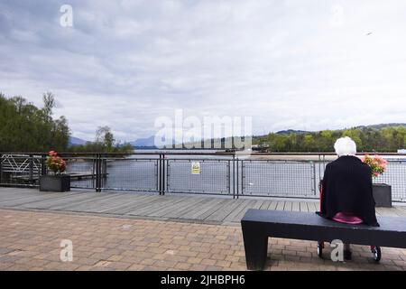 Elderly lady sits alone on bench looking out over Loch Lomond from Loch Lomond Shores Stock Photo