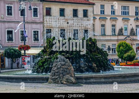 Banska Bystrica, Slovakia - August 17, 2021: view of fountain at SNP Square - stone base covered with green moss and water springs; historic buildings Stock Photo