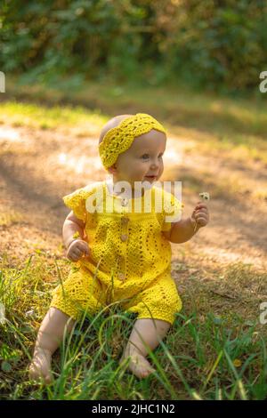 little cute happy baby girl sitting on the green grass on a sunny summer day dressed in knitted cotton yellow jumpsuit. Stock Photo