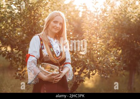 Young blonde woman in Serbian traditional holing a basket with fresh pears Stock Photo