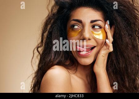 Enjoyed happy pretty Latin female with gold hydrogel patches under eye, stick tongue, looks up posing isolated over pastel beige background. Cosmetic Stock Photo